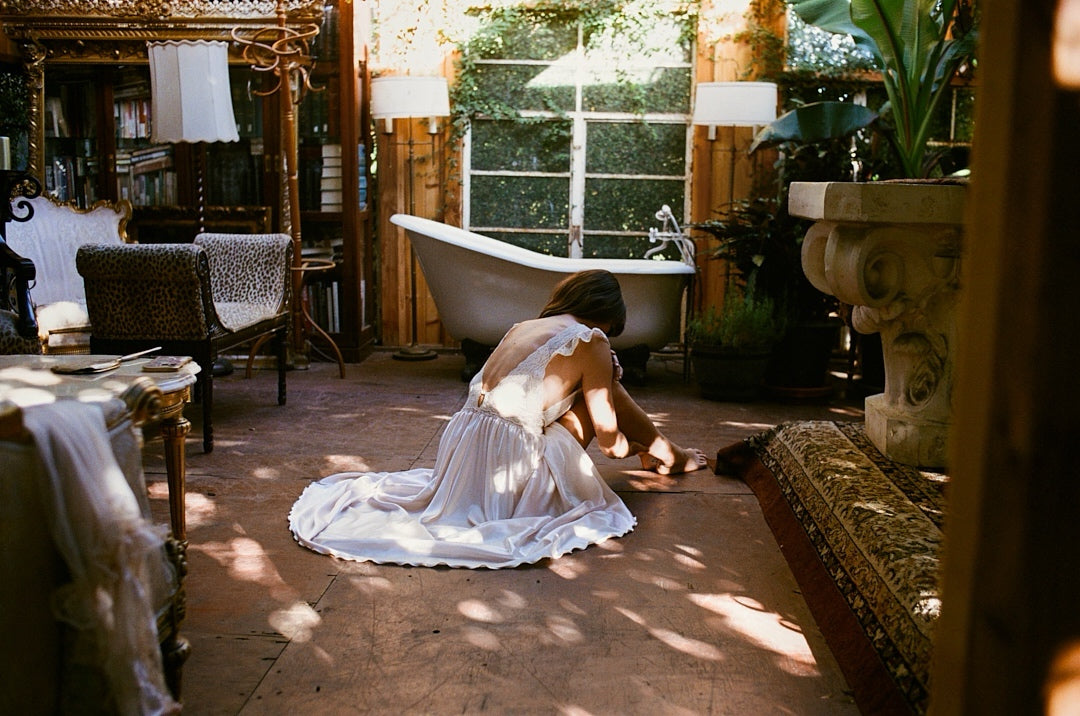 Woman in a white dress sitting on the floor in a sunlit room with a bathtub and decorative elements.